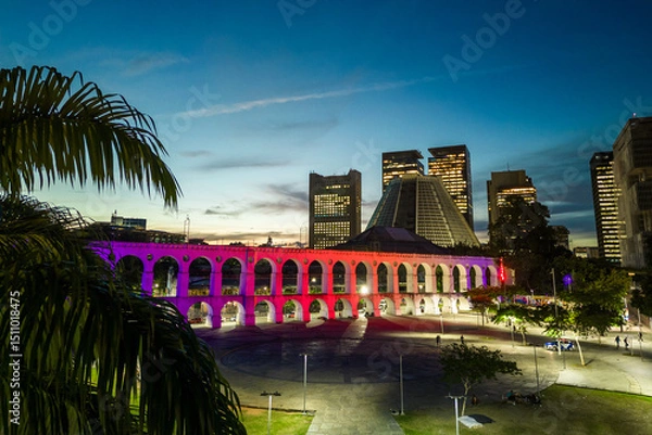 Fototapeta Lapa Aqueduct Illuminated With Purple and Pink Colors in the Evening With Tall Office Buildings of Rio de Janeiro Downtown