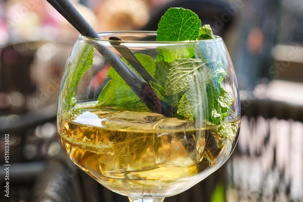 Fototapeta Cold summer drink or mocktail in a glass with ice cubes, fresh green mint leaves and a straw on a terrace in summer.