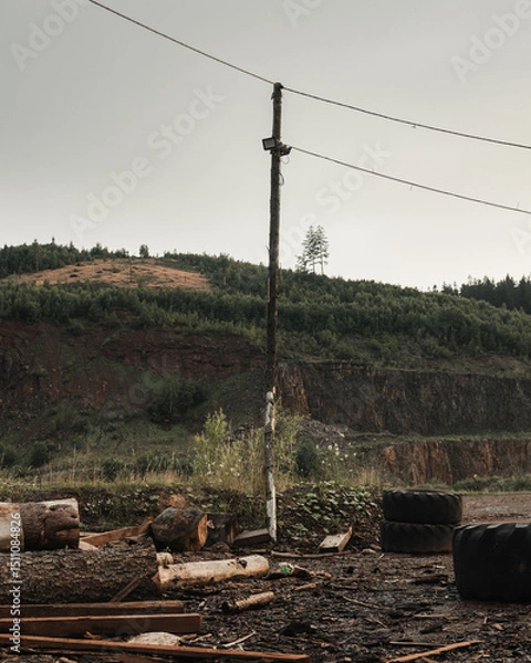 Fototapeta Abandoned Power Pole with Lines in the Raw Quarry Landscape with a Forest in the Background under the Cloudy Sky.