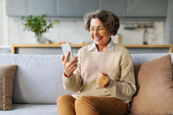 Fototapeta Happy elderly woman using smartphone at home, making video call, consulting doctor at virtual meeting on mobile phone and smiling at screen
