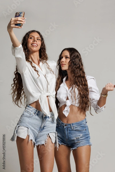 Fototapeta Two young women in casual summer outfits taking a selfie together against a neutral studio backdrop