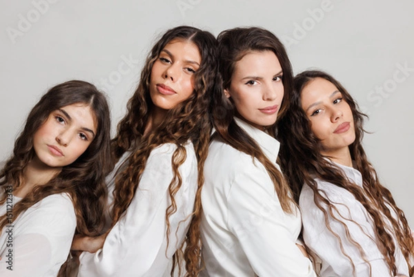 Fototapeta Four young women with long wavy hair posing together, dressed in white shirts, on a neutral background