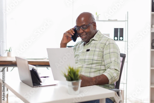Obraz Busy mature middle aged business man, senior professional executive manager talking on the phone making business call on cellphone sitting at work desk in office using laptop computer.
