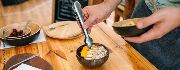 Fototapeta Banner of unrecognizable chef hands preparing a vegan breakfast made with mango, banana, raspberries, cereals and muesli using coconut bowl and metal tongs. Healthy vegetarian dishes concept.