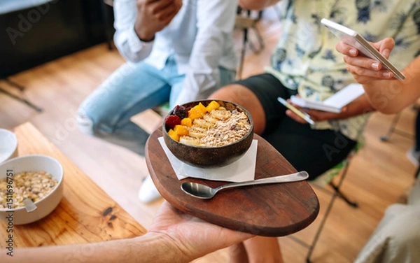 Fototapeta Unrecognizable chef showing vegan breakfast bowl made with mango, banana, raspberries, cereals and muesli to food bloggers and influencers taking pictures with smartphones in cooking master class.