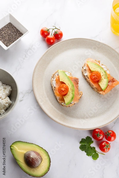 Obraz Top view of avocado, salmon and cherry tomato toasts on beige ceramic plate with ingredients around, on marble surface. Copy space. Concept of healthy breakfast and natural lifestyle