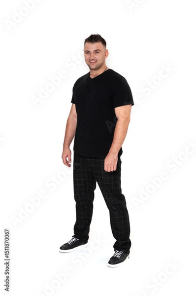Obraz A handsome dark-haired man in a black T-shirt, trousers and sneakers on a white background
