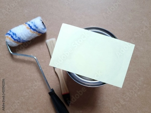 Obraz Blank sheet of paper on closed tin lid of paint can. Closed paint can and roller with brush on brown background