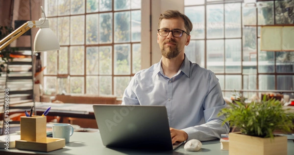 Fototapeta Bearded Professional Works on Laptop in Sunlit Office. Creative Man Looking at the Camera while Working from Home Office. Stylish Bright and Sunny Interior with Park in the Window. Medium Portrait