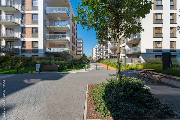 Fototapeta Wide pedestrian promenade in a new residential housing estate. This large public space includes various seating areas with benches and tables, surrounded by landscaped greenery and buildings.