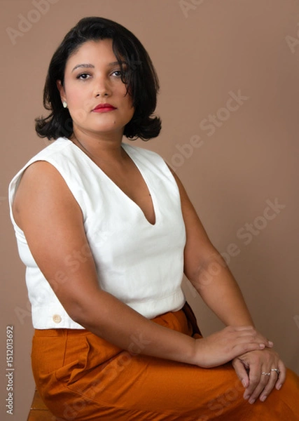 Fototapeta Elegant businesswoman in white top and orange pants sitting and looking at camera. Ideal for professional development, career, personal growth. Copy space on mocha mousse color background.