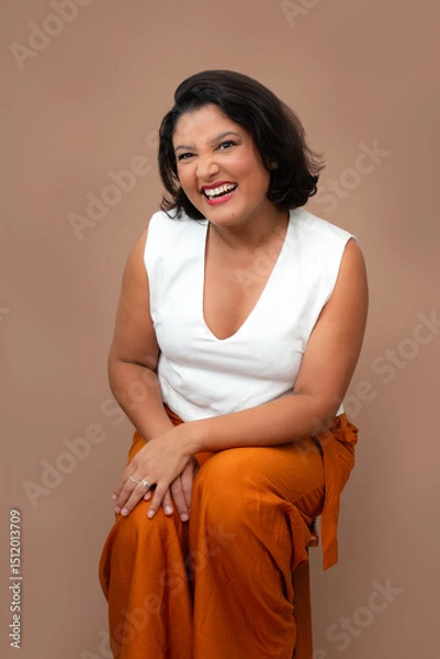 Fototapeta Cheerful businesswoman in white top and orange pants sitting on a stool and laughing at camera. Perfect for happiness at work, corporate well-being, friendly approach. Studio portrait with copy space.