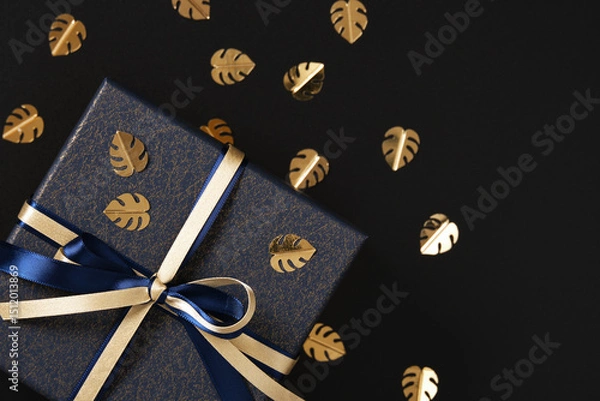 Obraz Fragment of blue gift box with gold and blue satin ribbon. Black background, gold confetti in the form of monstera leaf, space for text.