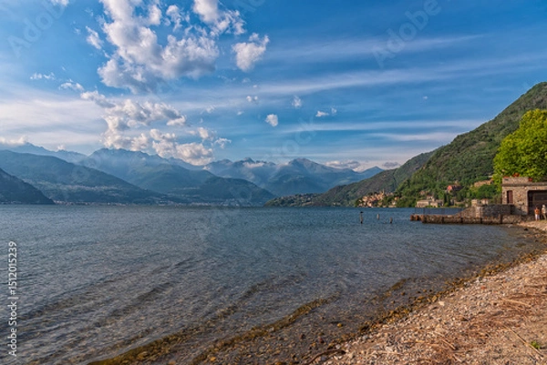Obraz A scenic lakeside village nestled at the foot of lush green hills, with majestic mountains rising in the background under a partly cloudy sky