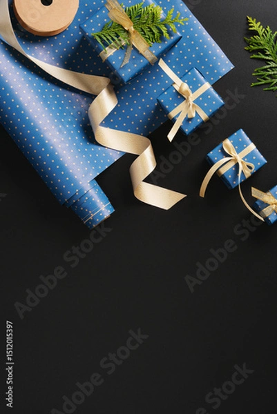 Obraz Flat composition with blue gifts with gold ribbons, brown paper tags, green thuja branches on black background. Space for your text.