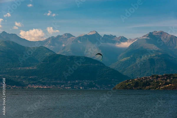 Obraz A scenic lakeside village nestled at the foot of lush green hills, with majestic mountains rising in the background under a partly cloudy sky