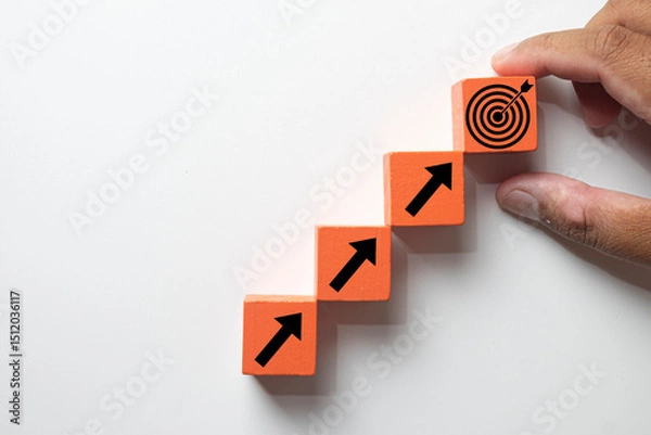 Obraz Hand placing an orange wooden block with a target icon atop ascending arrow blocks. Concept of business strategy, steps to achieving goals, progress, objective attainment, and success.