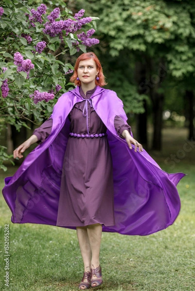 Fototapeta A beautiful young woman with red hair, against a background of blooming lilacs, in a cloak and hood. Medieval fantasy costume. Fairy tale heroine, princess, duchess.
