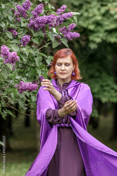 Fototapeta A beautiful young woman with red hair, against a background of blooming lilacs, in a cloak and hood. Medieval fantasy costume. Fairy tale heroine, princess, duchess.
