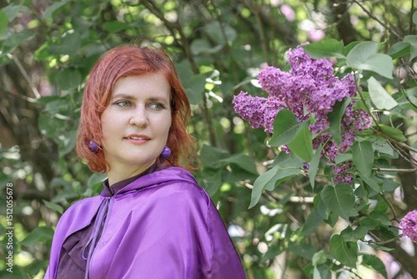 Fototapeta A beautiful young woman with red hair, against a background of blooming lilacs, in a cloak and hood. Medieval fantasy costume. Fairy tale heroine, princess, duchess.