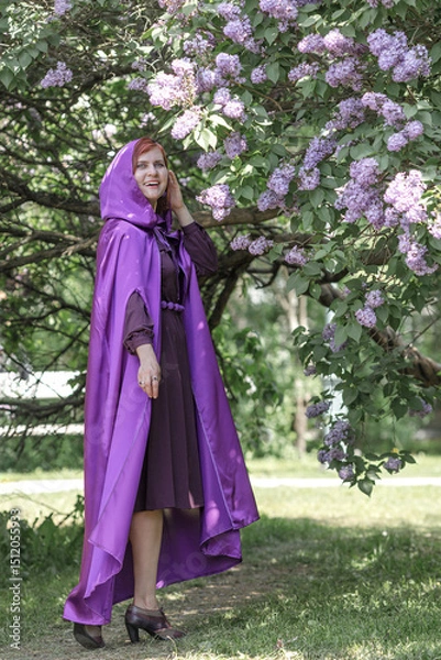 Fototapeta A beautiful young woman with red hair, against a background of blooming lilacs, in a cloak and hood. Medieval fantasy costume. Fairy tale heroine, princess, duchess.