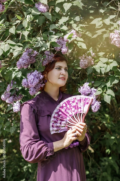 Fototapeta A beautiful young woman with red hair, against a background of blooming lilacs, in a cloak and hood. Medieval fantasy costume. Fairy tale heroine, princess, duchess.