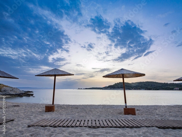 Fototapeta Tranquil beach at sunset with straw umbrellas overlooking the calm water and distant hills in Toroni on Sithonia peninsula, Greece