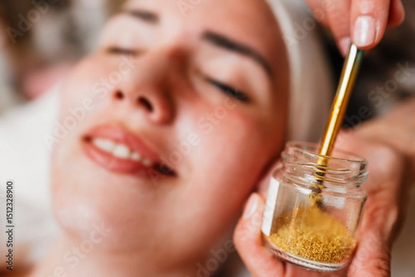 Obraz Beautician carefully applying gold particles with a brush on the forehead of a woman lying with closed eyes, during a rejuvenating treatment in a beauty spa, enhancing skincare and relaxation