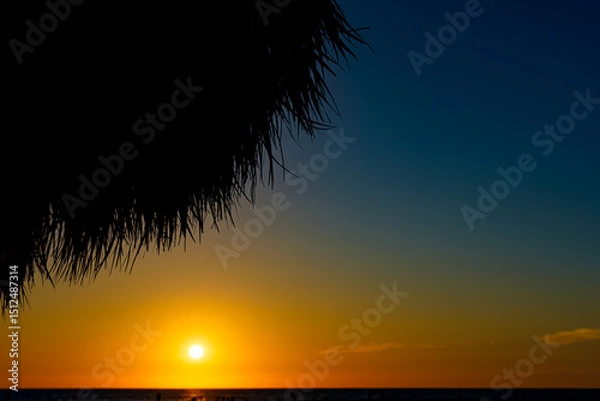 Obraz The sun sets on vacation, over the water while sitting on the beach, under an umbrella.