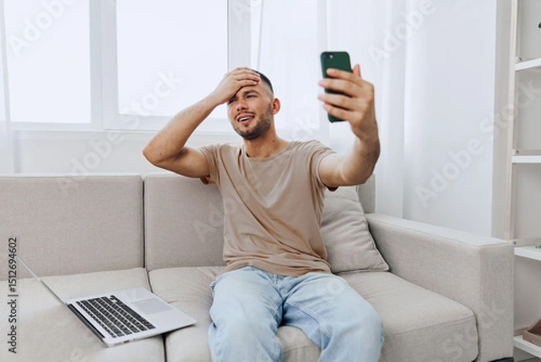 Fototapeta Smiling man sitting on a couch, excitedly video calling using a smartphone. Bright, modern living room with a laptop on the side adds to the cheerful atmosphere.