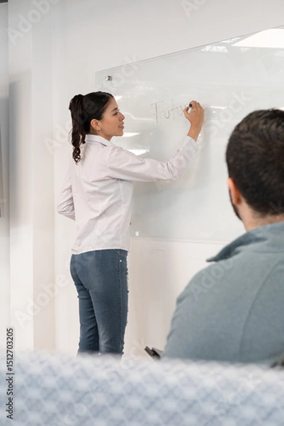Fototapeta Seen from behind, a businesswoman explains the project roadmap on a whiteboard during a strategic planning session in a modern office.
