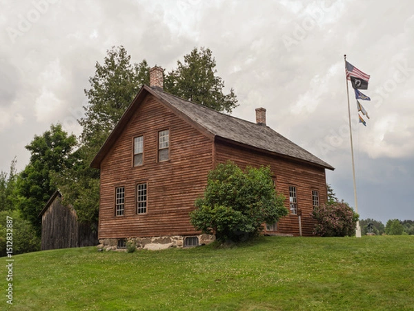 Obraz John Brown Farmhouse, with green lawn and waving American flag against cloudy sky in Lake Placid, New York State.
