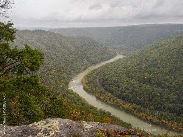Obraz Covered in early fall colors, the mountains at New River Gorge extend towards the horizon, framed by rocks in the foreground and drifting clouds above.
