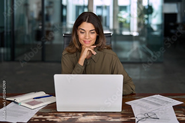 Obraz Smiling busy mature middle aged professional business woman, female company manager executive or entrepreneur looking at laptop computer in office working on online financial data sitting at desk.