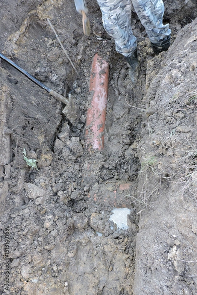 Fototapeta A worker wearing rubber boots standing in a deep ditch; a spade and a plastic orange pipe in marshy land next to a concrete cesspit to drain grounds