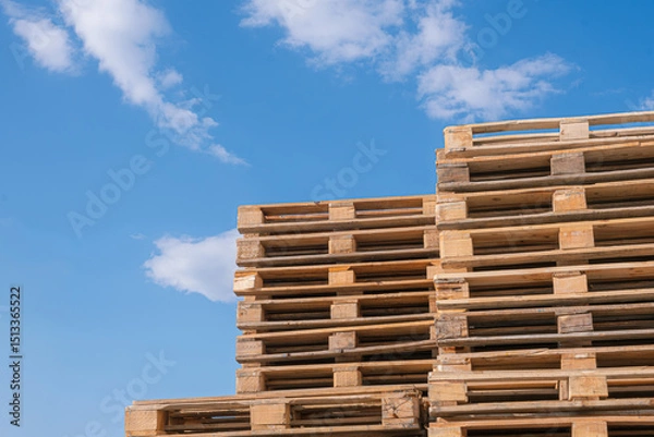 Obraz Stacked wooden pallets against blue sky. Large stack of wooden pallets arranged in an orderly manner. Related concept to logistics or warehousing.
