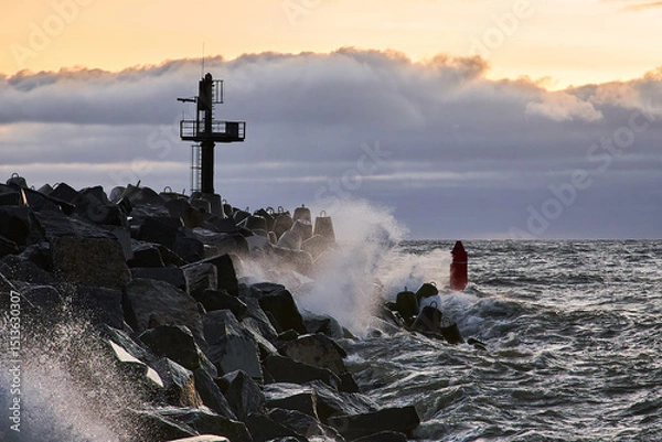Obraz The sea surf breaks on a stone breakwater against the background of sunset