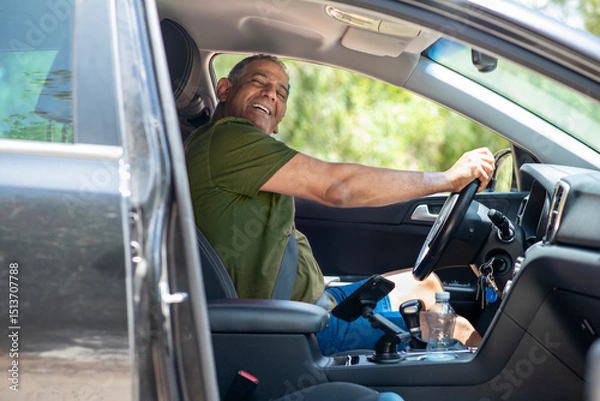 Obraz A middle-aged man smiles while sitting in the drivers seat of a car, enjoying a travel adventure. He appears relaxed and content, taking in the warm weather and scenic surroundings.