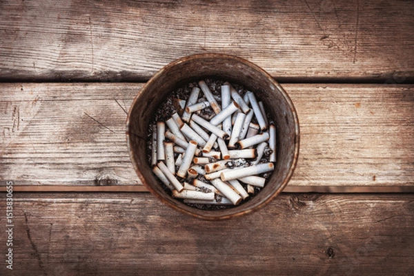 Fototapeta A metal container filled with numerous cigarette butts and ash sits on a weathered wooden surface, viewed from directly above. It's a stark photo of an unhealthy habit's aftermath.