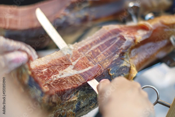 Fototapeta Close-up of traditional Spanish jamón being hand-sliced with a sharp knife. Thin slices of cured ham with rich marbling. Authentic gourmet delicacy in the making.