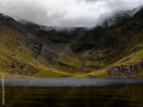 Fototapeta A tranquil mountain lake reflects the imposing cliffs shrouded in mist, evoking a sense of awe and solitude.