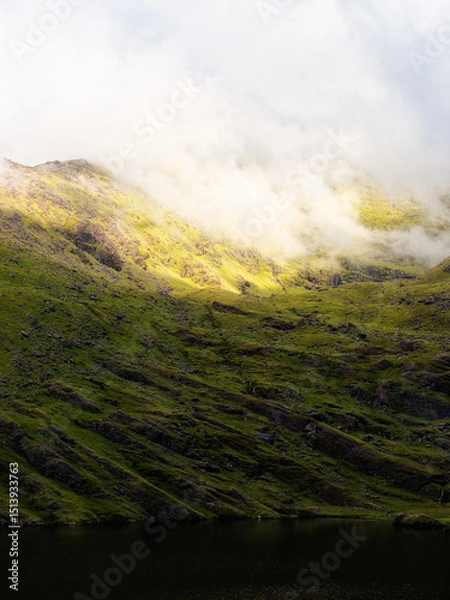 Fototapeta A grassy hillside slopes down towards the dark waters of Lough Callee. Clouds hover at the top of the hill, creating a serene and dramatic landscape in Ireland.