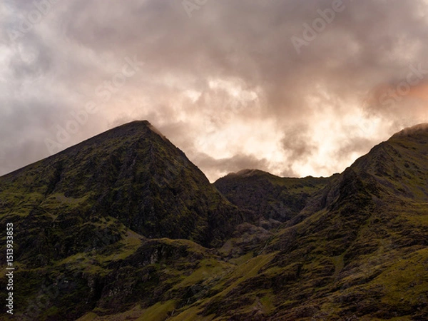 Fototapeta Carrauntoohil, the highest peak in Ireland, towers against a backdrop of swirling clouds as dusk approaches. The vibrant landscape displays lush green hills and rocky outcrops.