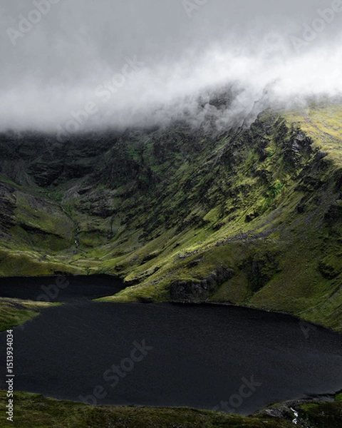 Fototapeta A mysterious mountain's peak is partially obscured by mist, its slopes meeting a dark, still lake. The image is evocative of solitude and grandeur.