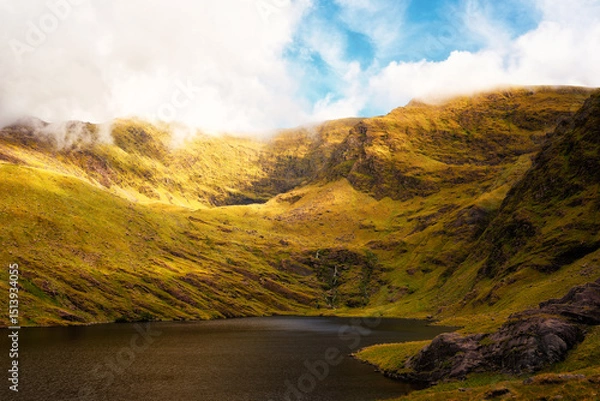 Fototapeta Scenic daytime showcases Lough Callee, nestled within green mountains in County Kerry, Ireland. Sunlight illuminates the mountainsides, with light clouds partially obscuring the peaks.