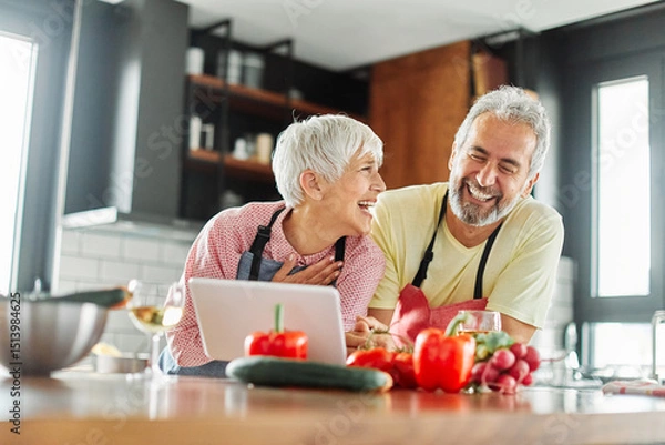 Fototapeta Portrait of happy senior mid aged mature couple prepering meal with fresh vegatebles and following internet instructions for a recipe on a tablet computer  or looking at video  or website app in kitch