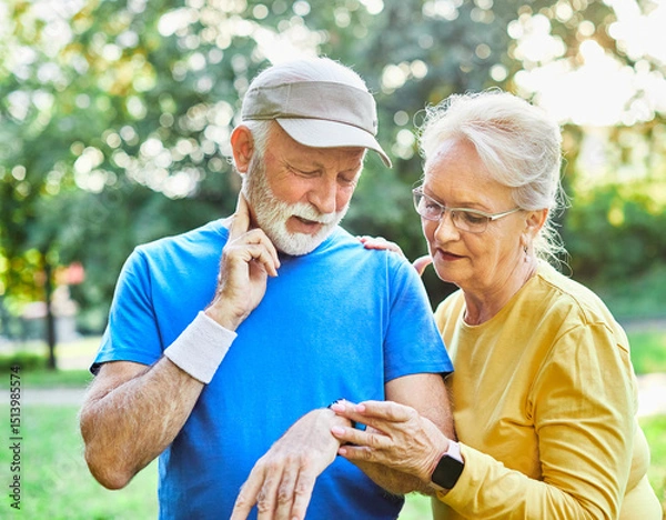 Fototapeta Smiling active senior couple jogging exercising and having fun and laughing together taking a break in the park, senior athletes checking their er heart rate with smartwatch
