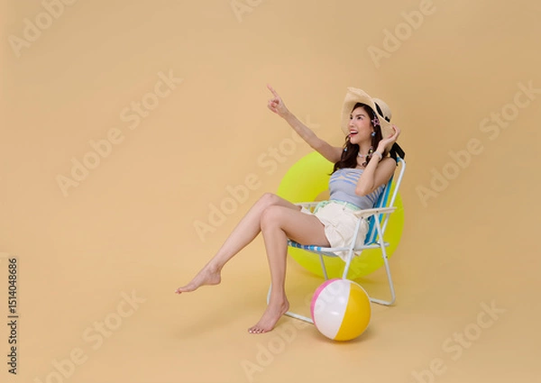 Fototapeta Happy Asian woman dressed in summer clothes sitting on beach chair and pointing finger isolated on nude background. Summer vacation sea beach rest concept.