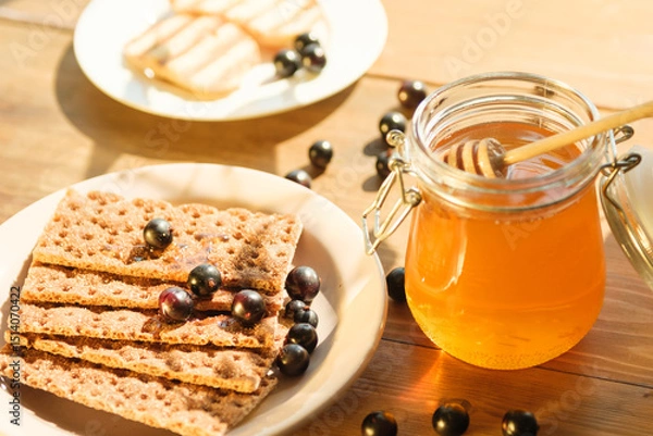 Fototapeta Fresh honey in a jar with a dipper rests next to crunchy crackers and berries on a rustic wooden table, illuminated by warm sunlight, creating an inviting atmosphere.