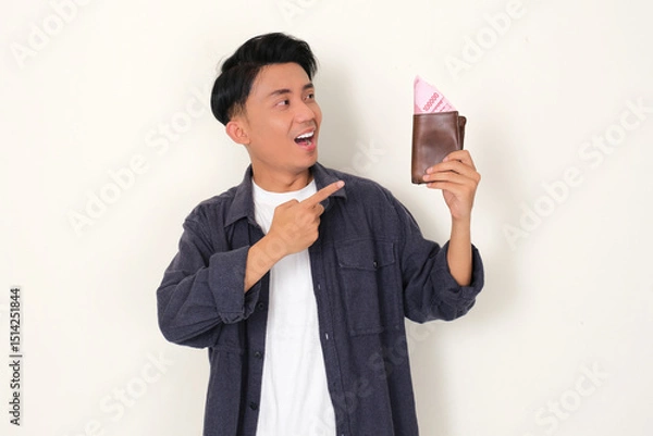 Fototapeta Young Asian man showing happy face while looking and pointing at wallet full of money that he hold